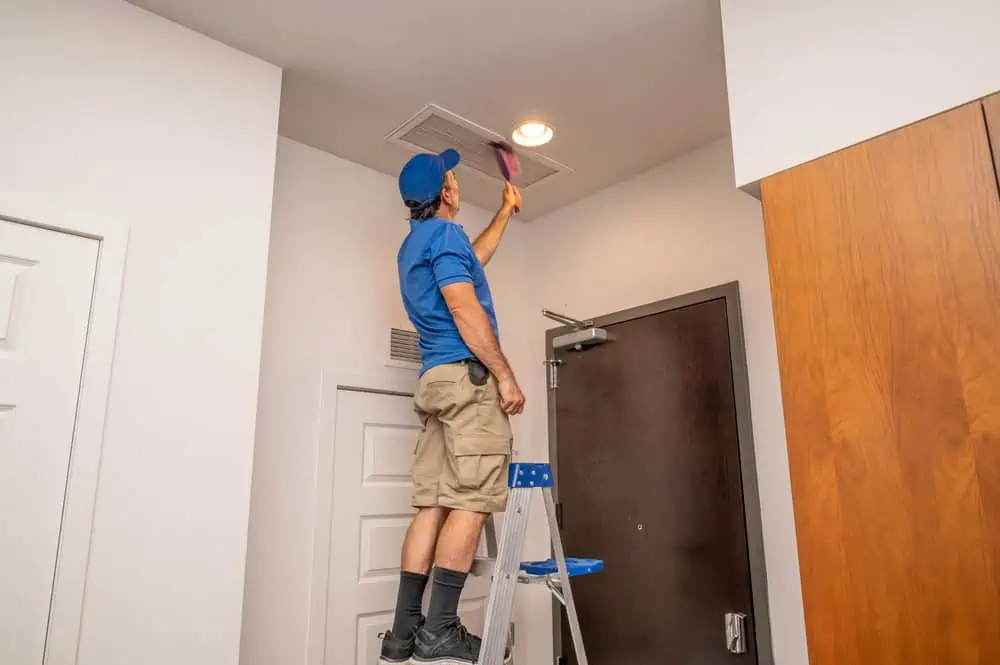 A person in a blue shirt and cap stands on a ladder cleaning an air vent on the ceiling with a brush in a room with white walls and a wooden door.