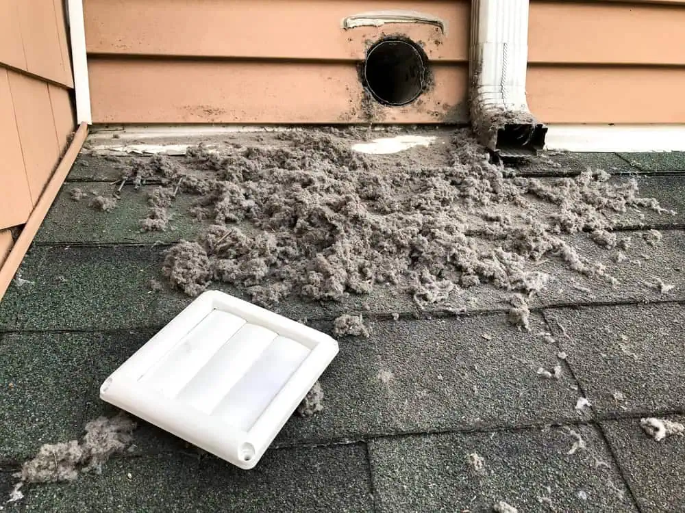 Ejected dryer lint scattered on a dark green roof beneath a broken white vent cover, with an exposed vent and a nearby gutter downspout visible on the side of a house.