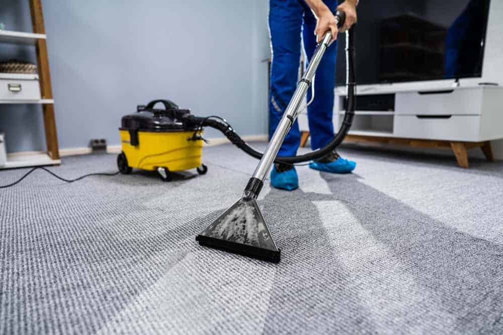 A person uses a yellow and black carpet cleaning machine to steam clean a gray carpet in a modern living room. The focus is on the cleaning process and equipment.
