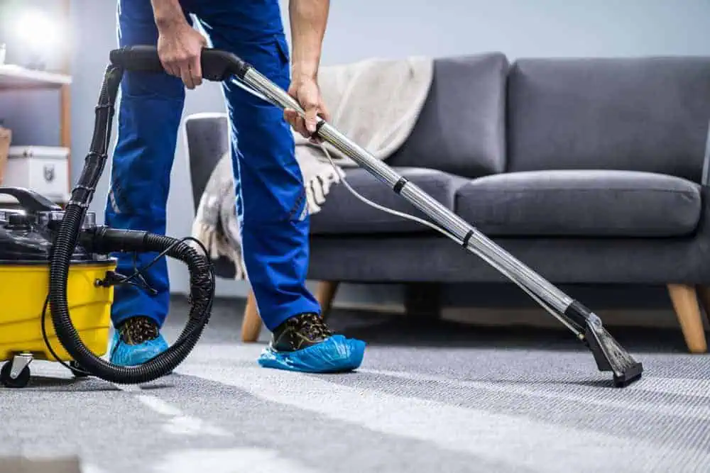 A person wearing blue coveralls and shoe covers uses a vacuum cleaner on a carpeted floor in a living room with a gray sofa and a blanket in the background.