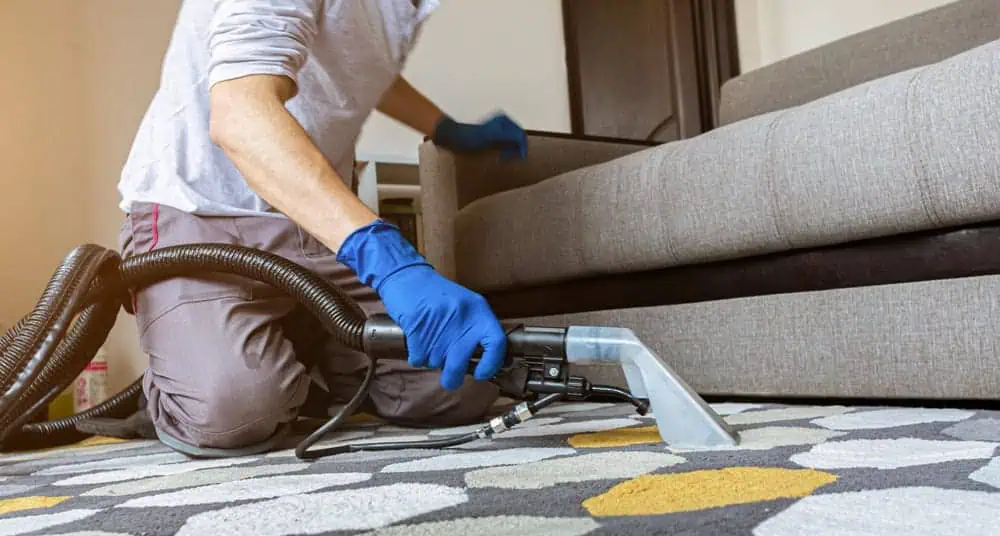A person wearing blue gloves uses a carpet cleaning machine on a patterned rug in a living room, kneeling beside a gray sofa.