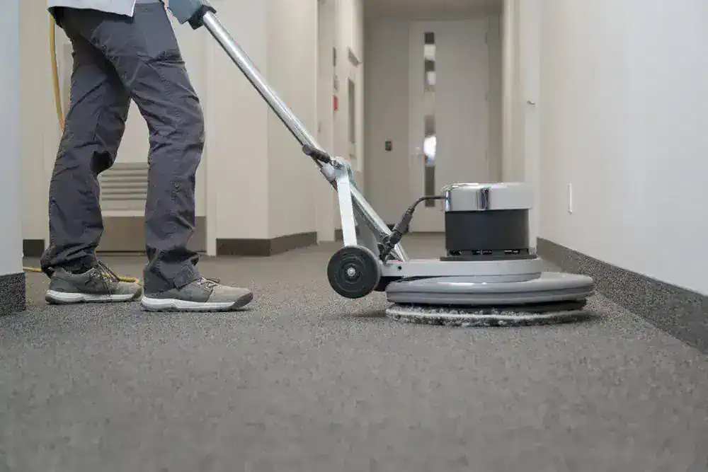 A person is using a large floor cleaning machine to clean the carpet in a hallway with light-colored walls and doors. Only the lower half of the person is visible.