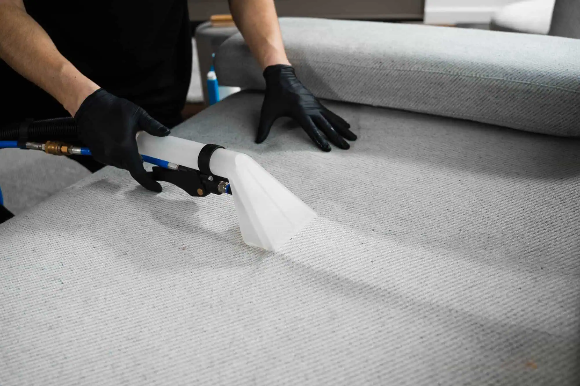 Person wearing black gloves using a professional upholstery cleaning tool to clean a light gray fabric sofa. The scene focuses on the cleaning process, showing the texture of the fabric and the cleaning equipment.