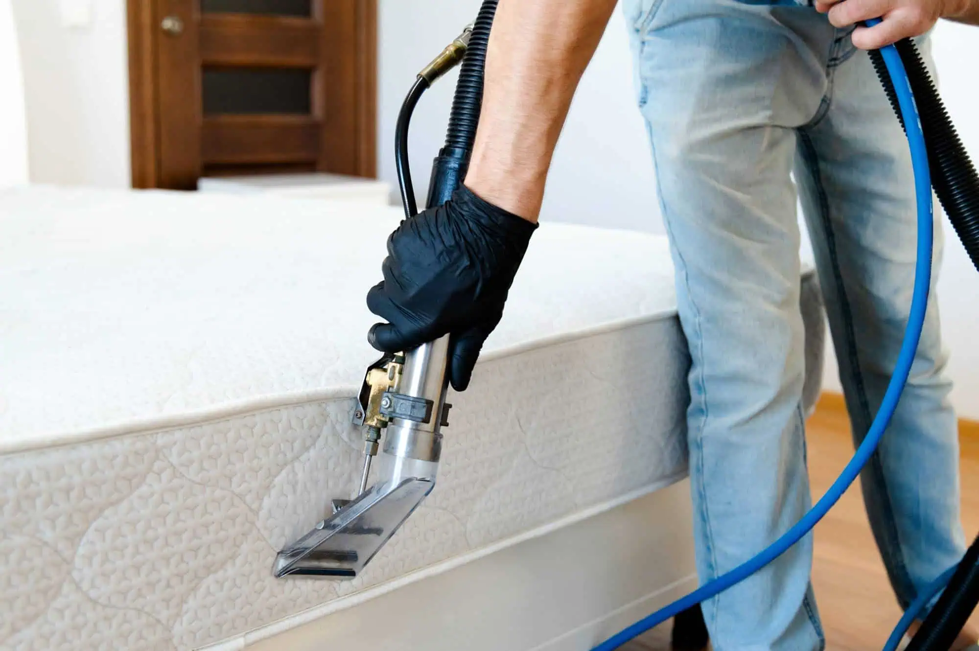 A person wearing black gloves uses a professional steam cleaning tool to clean a mattress in a bright room with wooden floors and a brown door in the background.