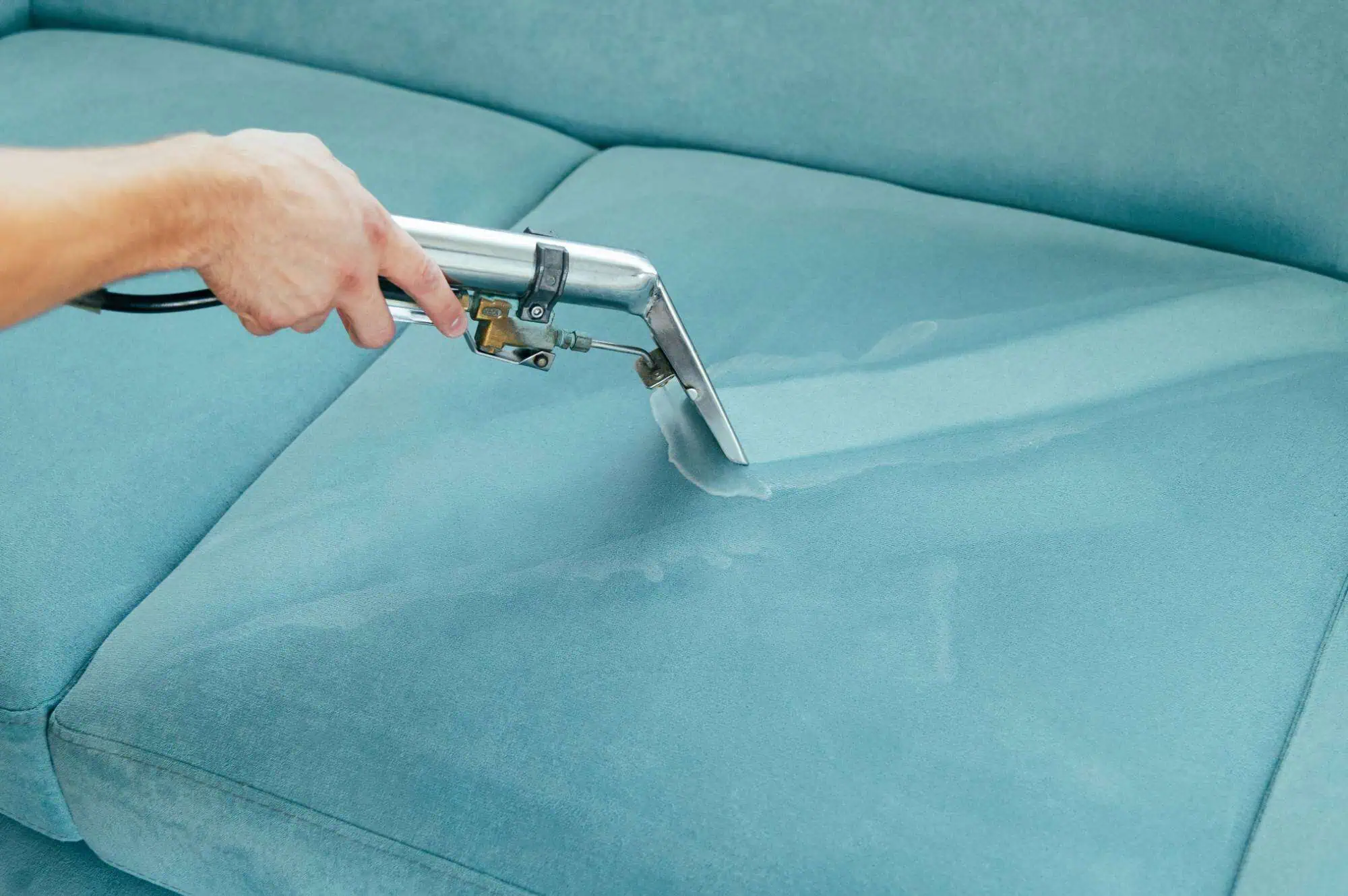 A person uses a handheld upholstery cleaner to clean a light blue fabric sofa, visibly removing dirt and moisture from the surface.