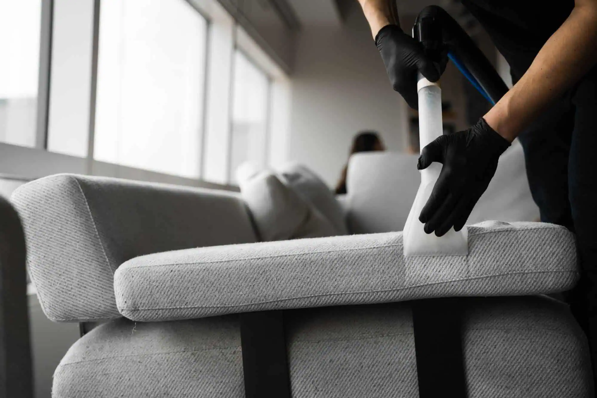 A person wearing black gloves uses a vacuum cleaning tool to clean a light gray couch cushion in a brightly lit room with large windows.